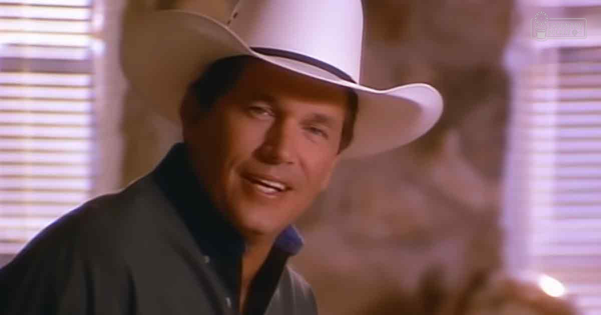 A middle-aged adult male wearing a dark-colored, collared, regular-fit jacket with long sleeves and a white cowboy hat stands indoors against a softly lit, rustic or Western-themed backdrop.