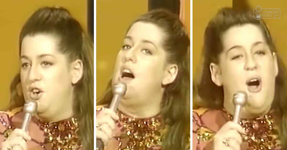 A young adult female wearing a sparkly gold and red sequined top sings into a microphone against a plain yellow background, suggesting an indoor stage setting.