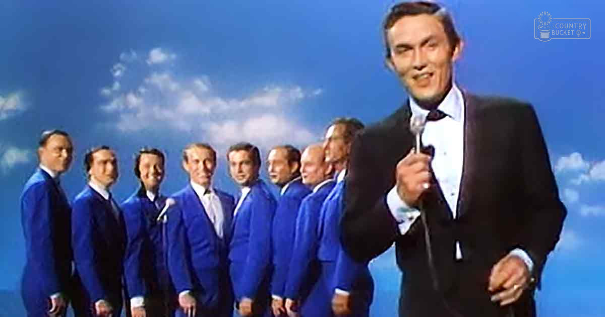 A group of men in blue suits stands against a stage backdrop with blue lighting and a sky-like design.