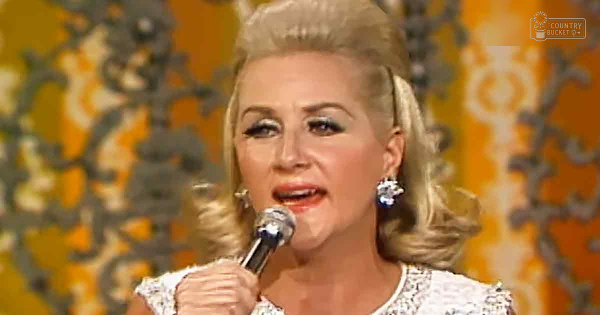 A middle-aged woman in a white lace, high-necked vintage-style outfit with large dangling earrings performs on stage against ornate wallpaper.