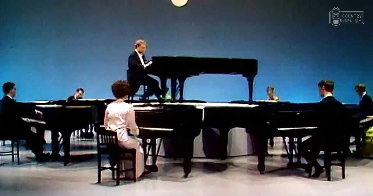 A middle-aged adult male pianist in a dark suit jacket and trousers performing with other musicians, including a young adult female performer in a light-colored dress or blouse with a patterned skirt or pants.