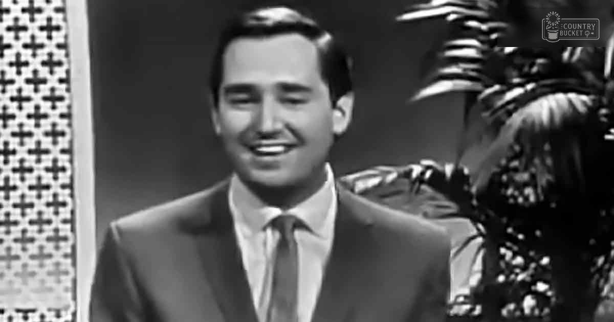 A young adult male wearing a mid-20th century suit jacket, white dress shirt, and dark tie stands indoors with decorative elements in the background.