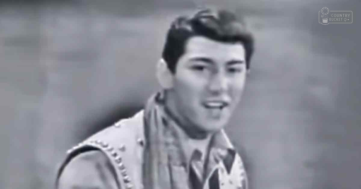 A young adult male wearing a fitted sleeveless vest with decorative patterns over a collared shirt in an indoor setting.