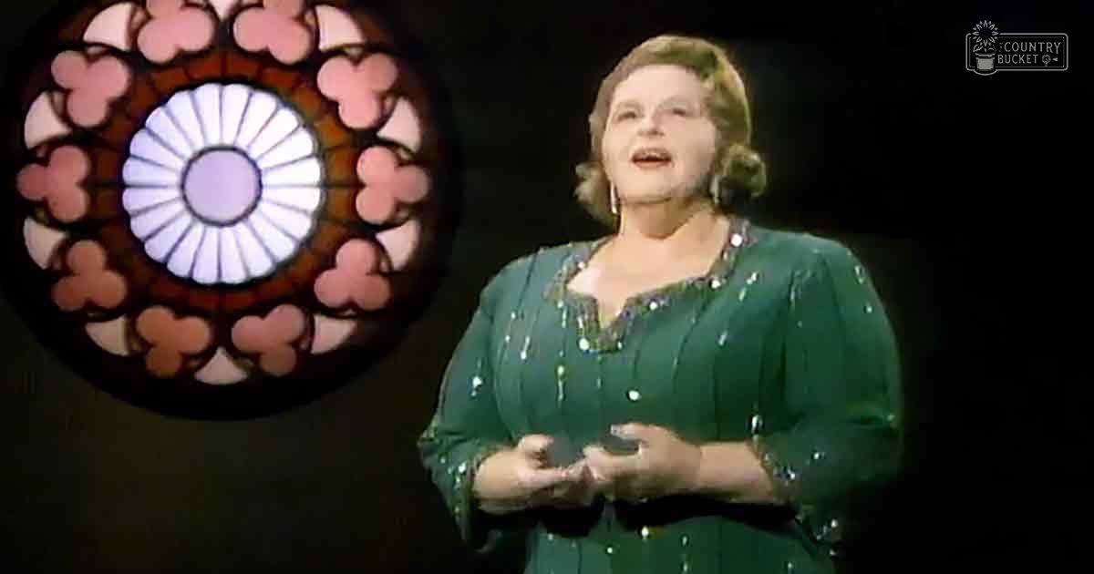 An older adult female singer wearing a green sequined or beaded dress with long sleeves, performing indoors at a stage with dark lighting and stained glass window behind her.