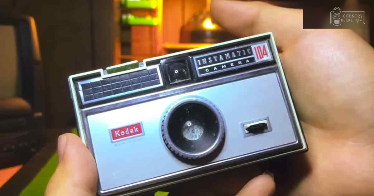 A Kodak Instamatic 104 camera held indoors with warm lighting and wooden furniture in the background.