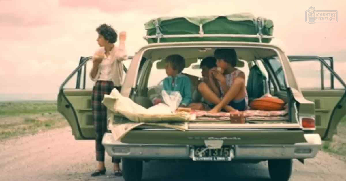 A family scene in an open landscape with clear skies and sparse vegetation, with a vehicle parked on a dirt road suggesting travel or camping.