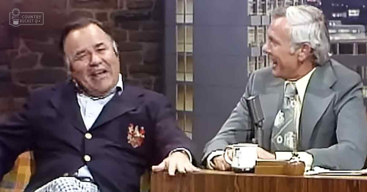 A talk show set with two middle-aged men in formal attire seated at a wooden desk under natural light from large windows.