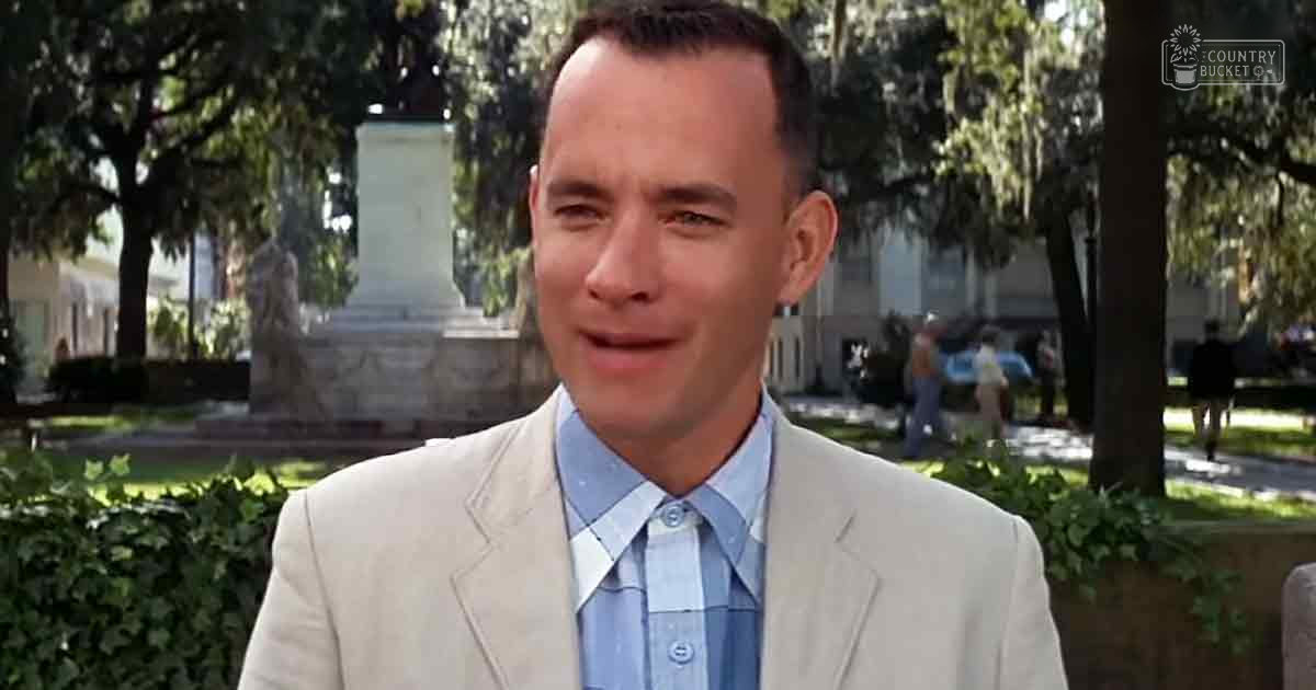 A middle-aged man wearing a light beige suit jacket over a light blue dress shirt and tie stands outdoors near trees and a stone monument, with greenery and people walking in the distance.