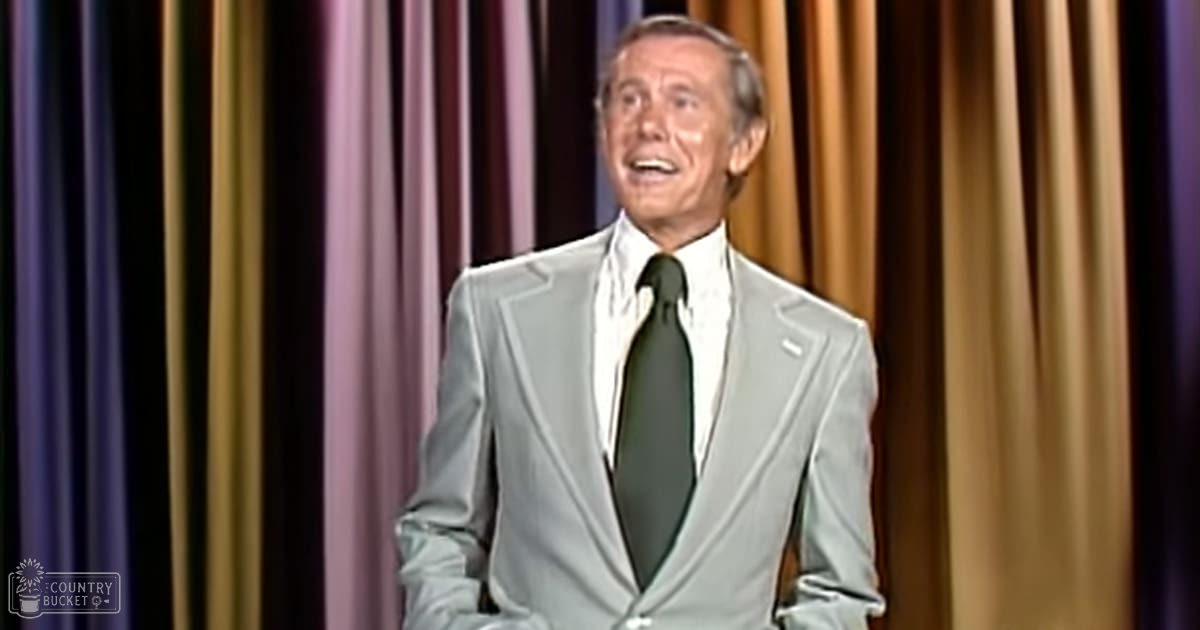 A middle-aged man in a light gray suit jacket, white shirt, and dark green tie stands indoors with purple, gold, and brown vertical curtains suggesting a stage or performance setting.