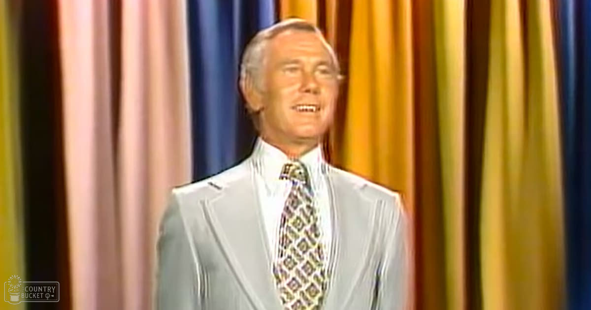 Older adult male in a studio setting with light gray suit jacket with subtle pattern, white dress shirt, and geometric tie; vertical curtains of yellow, blue, pink, and brown in the background.