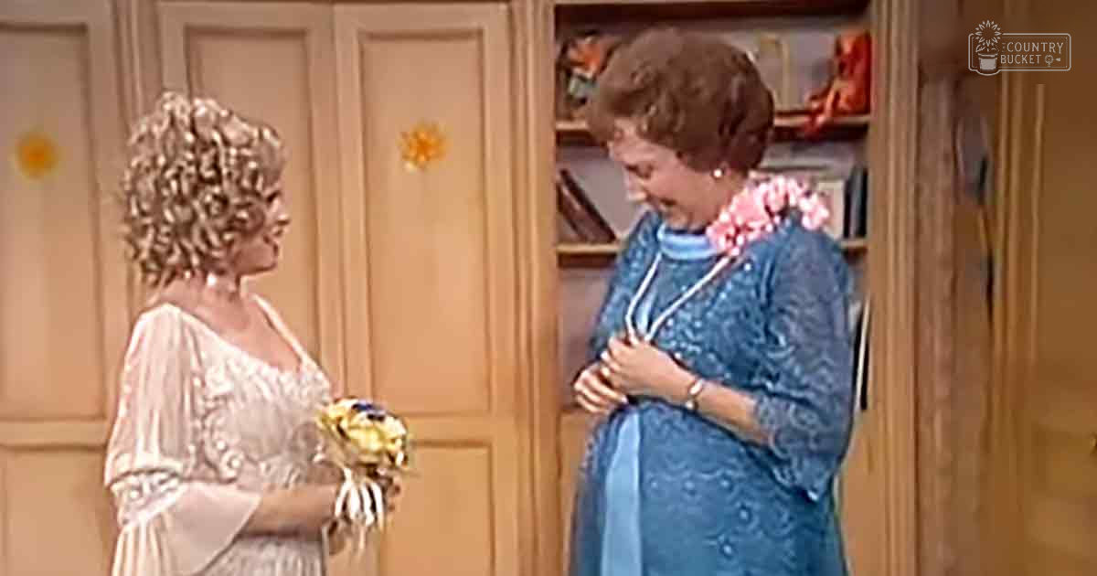 A home interior scene with two adults, one wearing a light-colored dress holding flowers, another in a blue floral-patterned dress adorned with jewelry; furniture and decor elements are visible.
