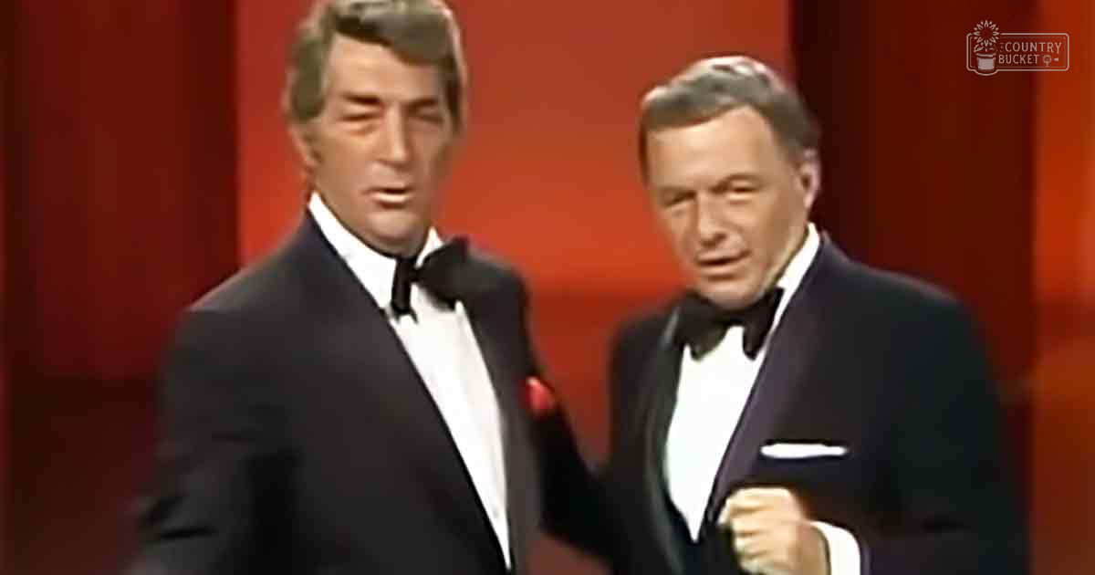 Two adult men in a formal setting, possibly on stage or in a studio. They wear black tuxedos with distinct details from the mid-20th century, against a red backdrop and professional lighting.
