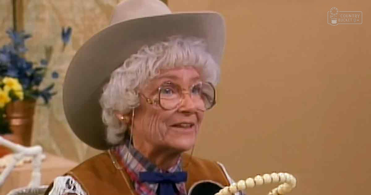 An older adult woman wearing a wide-brimmed hat, glasses with thick frames, and a patterned scarf in a domestic setting with yellow and blue flowers and beige tones.