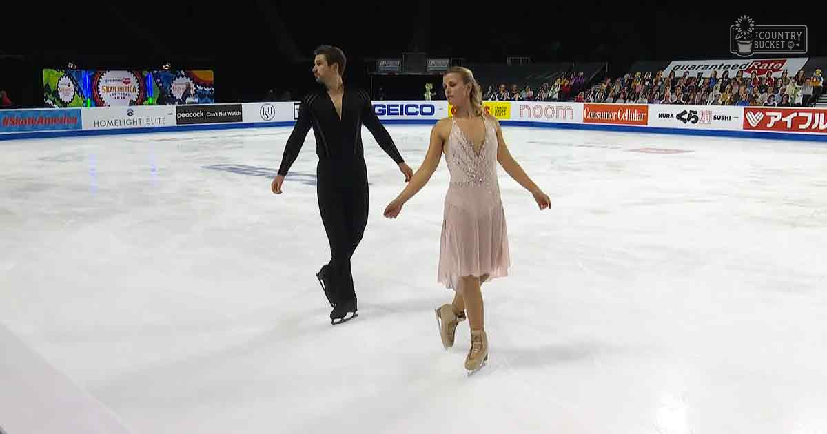 Figure skating pair performing at an indoor ice rink event with banners of sponsors visible, audience seated in the stands under artificial lighting suggesting evening or night time.