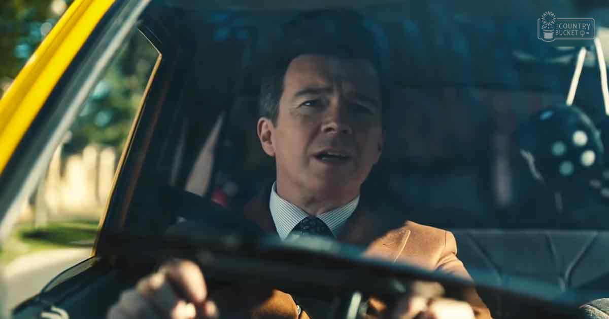 An adult male wearing a light brown suit jacket with a white dress shirt and dark patterned tie, driving an older model yellow car outdoors.