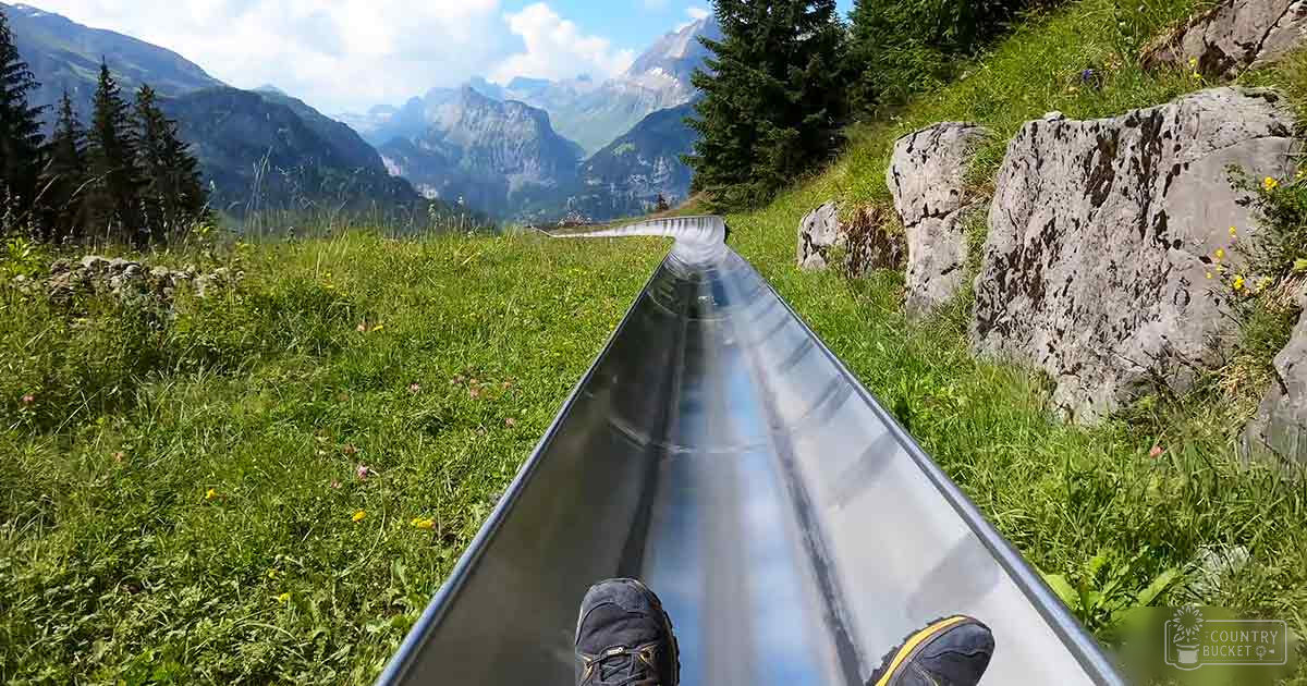 An outdoor alpine slide scene with grassy fields and rocky terrain under a partly cloudy sky.