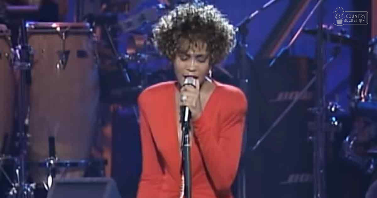 An adult female singer in a red 1980s dress singing into a microphone on a stage set with musical instruments and professional lighting.
