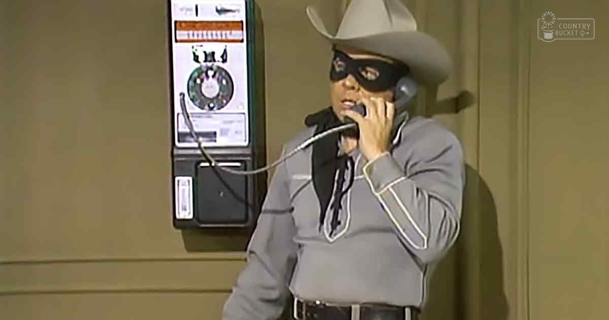 An adult male wearing a gray suit with white stripes, black shirt, and wide-brimmed cowboy hat stands indoors near an older model payphone labeled 'Madly Odd'.