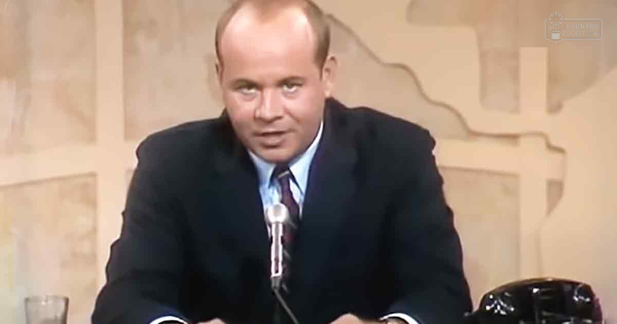 A middle-aged adult male wearing a dark suit jacket with a patterned tie, light blue collared shirt, and high-quality fabric in an indoor professional setting.