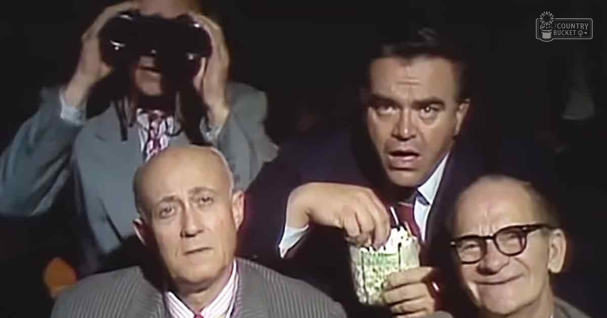A man in the foreground holding popcorn at an indoor event with dim lighting, likely evening or night time.