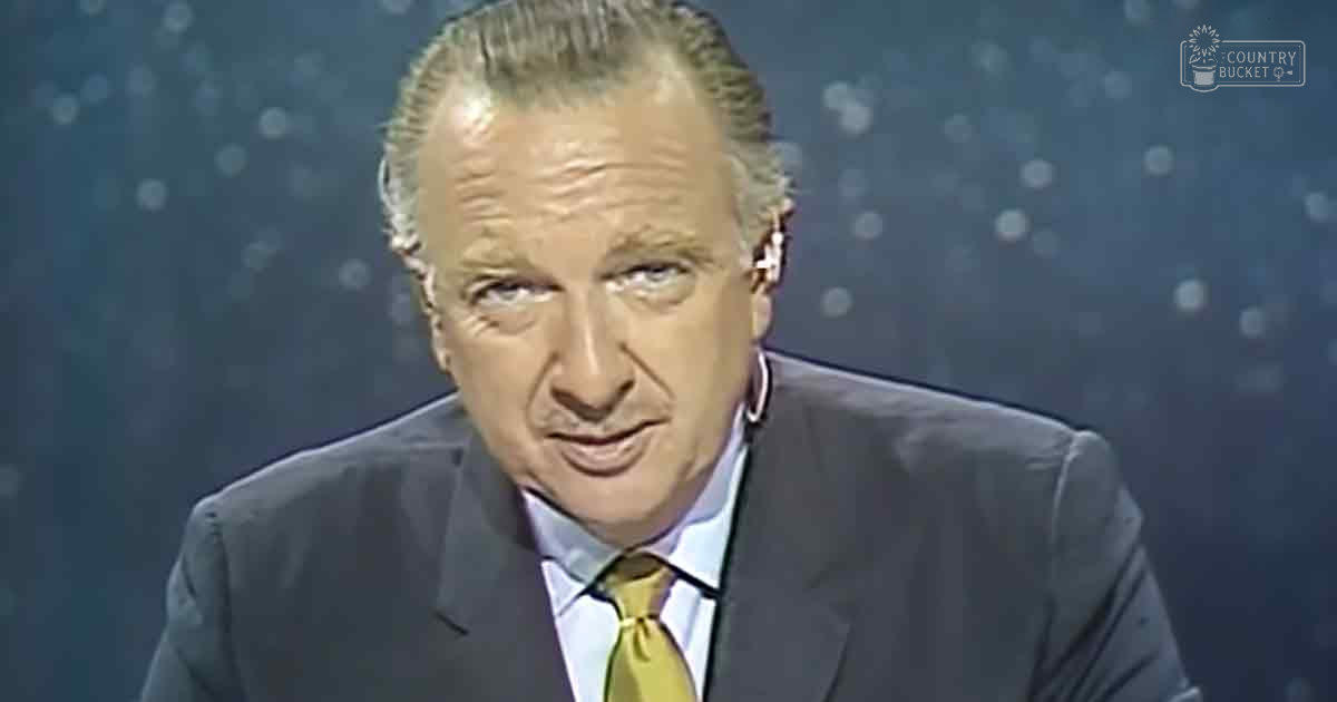 An older adult male in a studio setting with dark suit, light blue dress shirt, gold tie against a dark blue backdrop with bokeh lights.