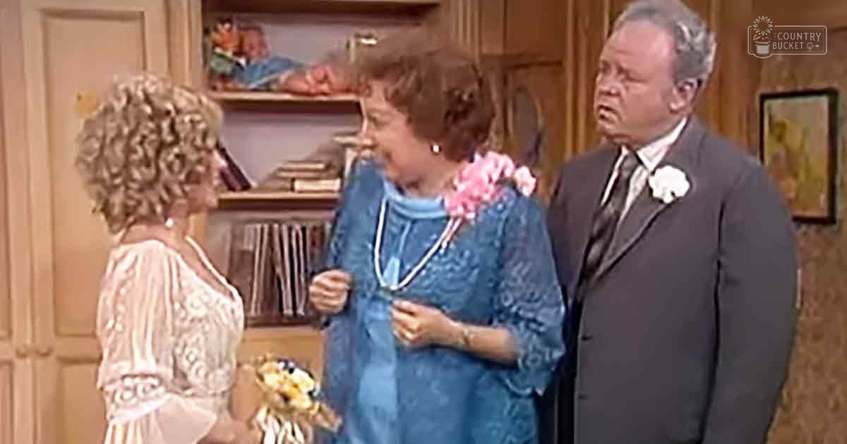 A middle-aged woman wearing a white lace dress with long sleeves in an elegant flowy style, holding yellow flowers; another middle-aged woman in a blue floral-patterned dress and necklace with pendant, also holding pink flowers in her hair; a middle-aged man in a dark gray suit jacket and tie, standing in a domestic living room or dining area with wooden bookshelves and framed pictures on the wall.