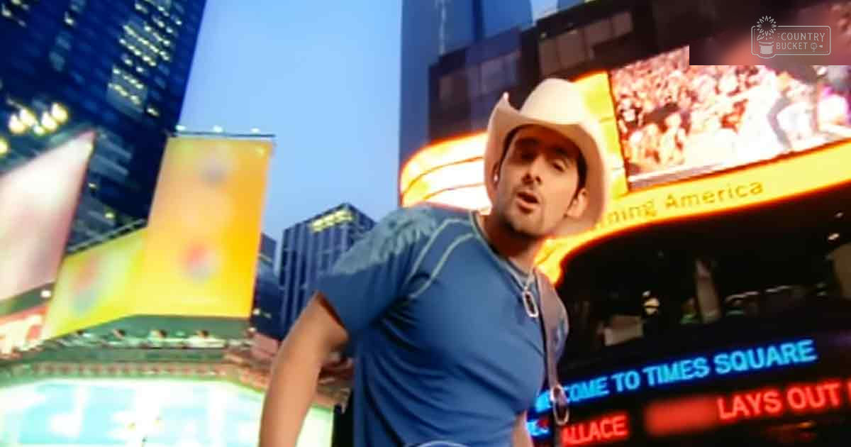 A young adult male wearing a blue t-shirt with stars and a white cowboy hat stands on Times Square at night, surrounded by bright lights and advertisements.