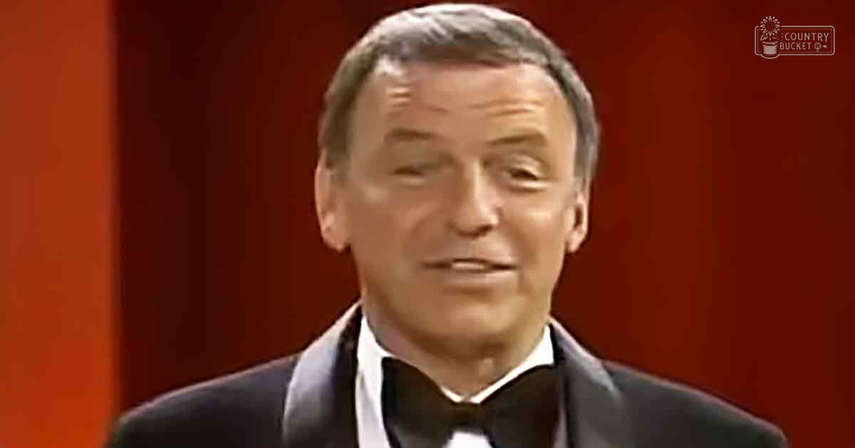 An older adult male wearing a classic fit tuxedo with a collared shirt, standing against a red backdrop in what seems to be an indoor performance or broadcast setting.