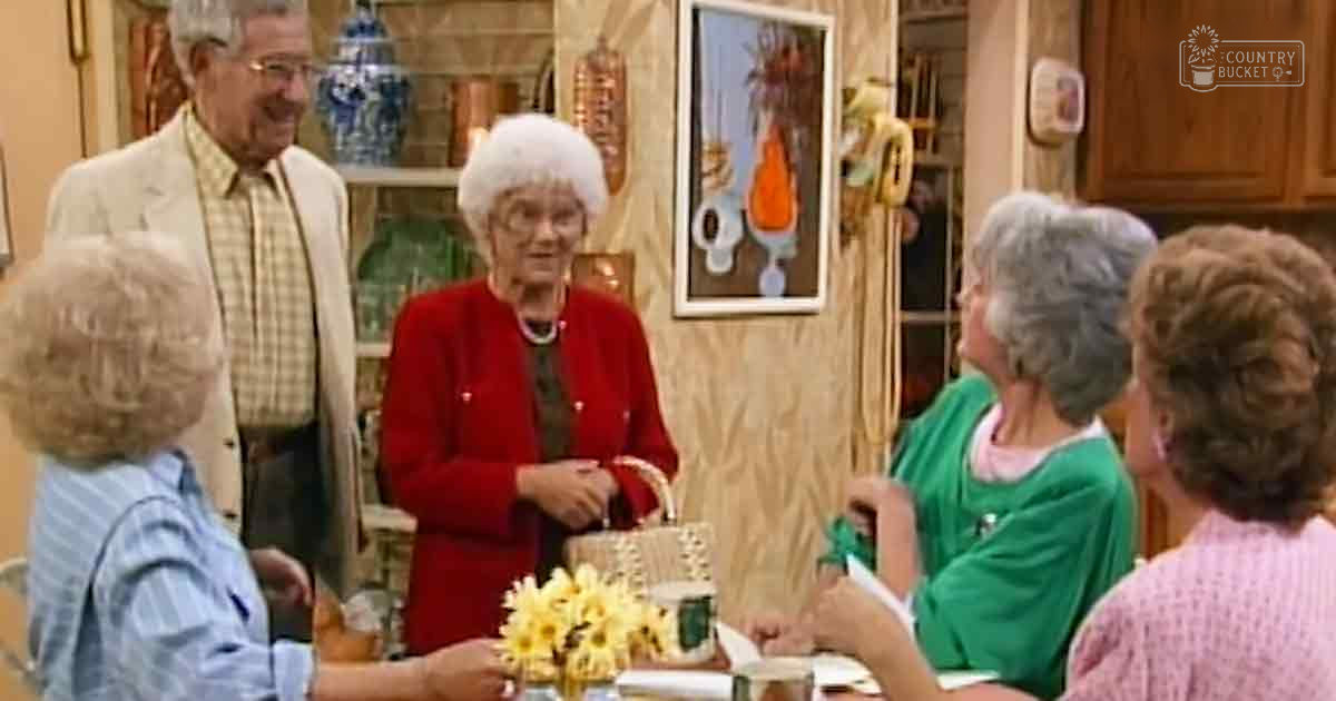 An older adult woman wearing a red dress with black belt stands at a dining table in a home setting, surrounded by other adults engaged in conversation; wall decor and natural light suggest daytime.