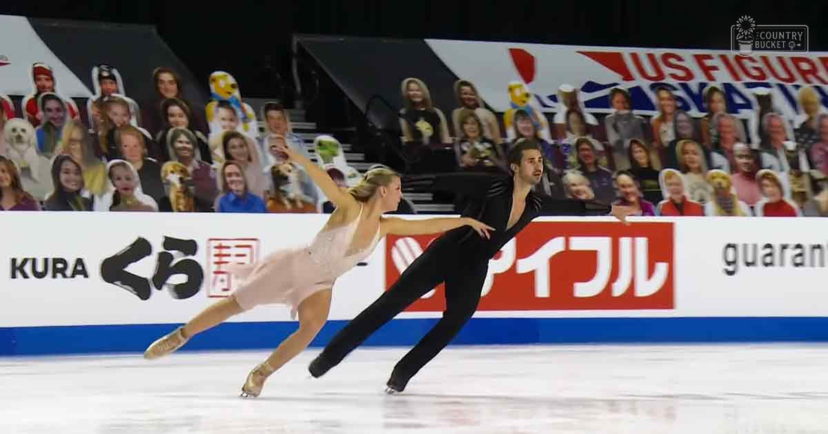 A figure skating pair performing at an indoor ice rink, with a young adult female in a light pink satin dress and a young adult male in a black form-fitting jumpsuit. Banners display 'US Figure,' 'KURA くら,' and 'guaran.'