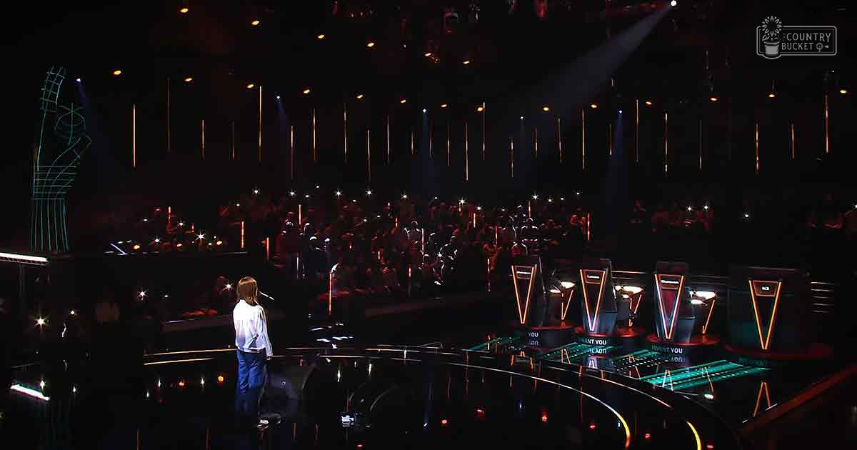 A young adult female performer stands on stage at an awards ceremony, wearing a white shirt and blue jeans under bright stage lights with an audience holding up lights in the background.