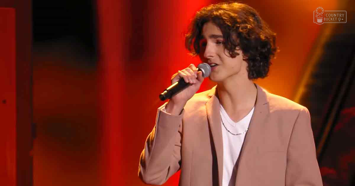 A young adult male singer performing on a stage bathed in red lighting, wearing a light brown blazer over a white t-shirt.
