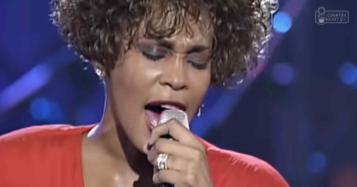 An adult female performer singing into a microphone, wearing an orange dress with large hoop earrings and multiple rings; studio setting with bokeh lights.