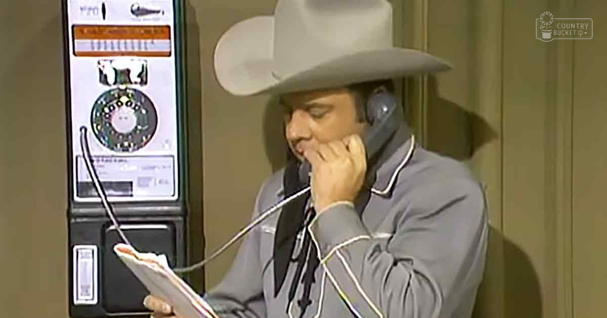 An adult male in a relaxed fit, collared gray Western-style shirt with long sleeves and a wide-brimmed cowboy hat stands indoors, holding what looks like paper or script while talking into a vintage payphone. The setting is likely a professional studio or office environment.