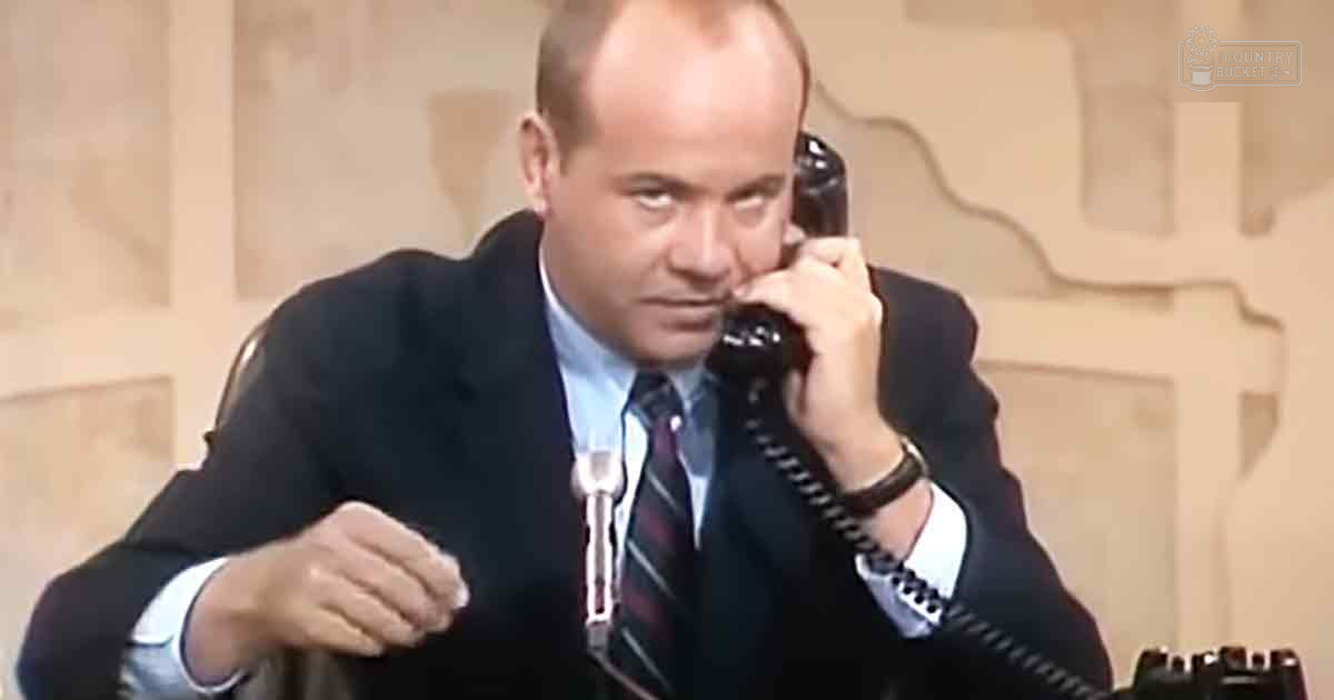 A professional adult male wearing a dark suit with a striped tie, sitting at an office desk holding a black rotary phone.