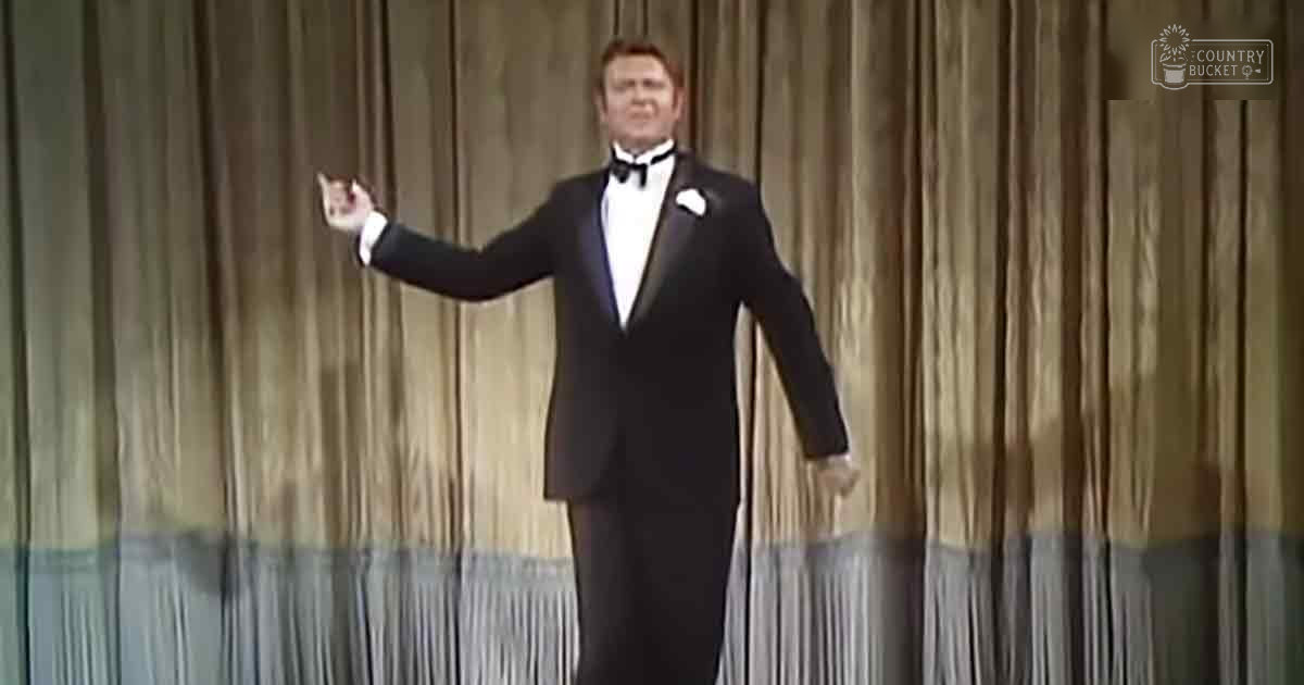 A formally dressed adult male performer in a mid-20th century tuxedo suit, complete with white shirt and black bow tie, stands on an indoor stage set against curtains.