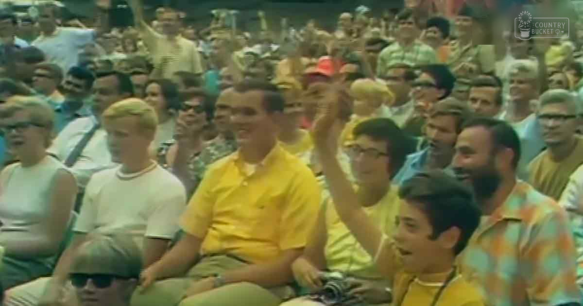 A daytime outdoor stadium or arena filled with spectators, possibly a sports game or concert; children and adults in casual summer attire under natural light.