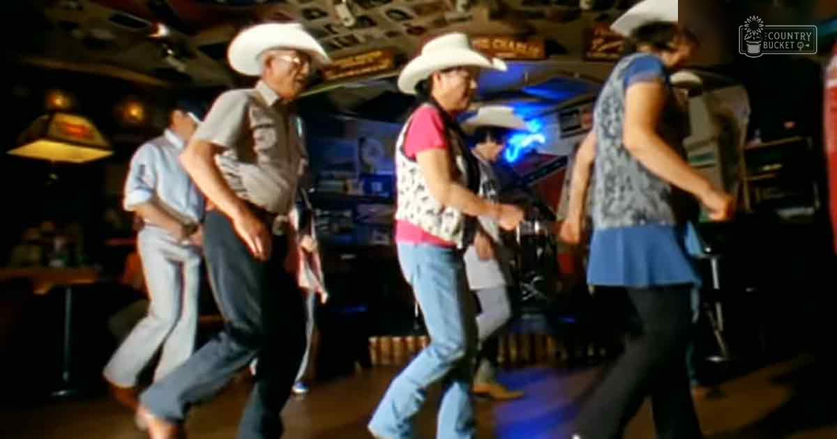 Three older adults dancing in a dimly lit bar with wooden furniture, signs reading 'Champ', and casual attire with cowboy hats and light blue shirts.