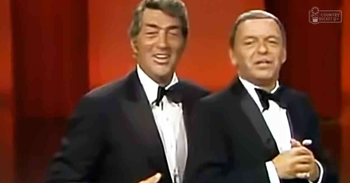 Two adult males wearing formal black tuxedos with white shirts and black bow ties, standing against a backdrop of red curtains in an indoor setting.