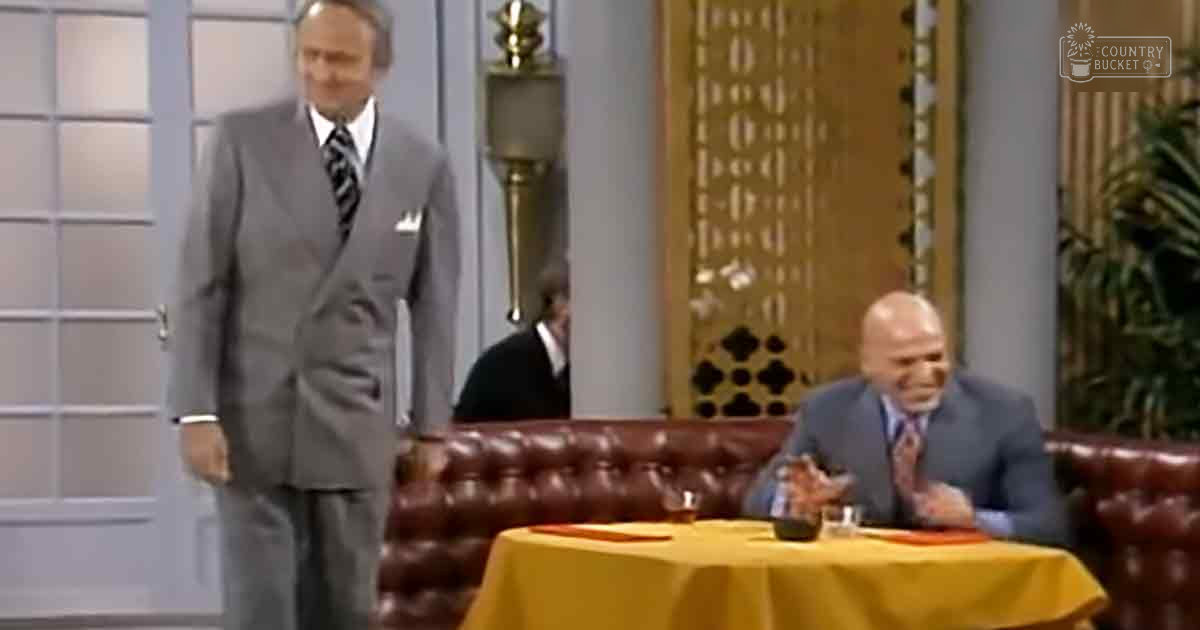 A middle-aged man wearing a gray suit and white shirt with patterned tie stands near an older adult male in a blue suit jacket, white shirt, and striped tie at a round table covered with a yellow tablecloth. The setting is indoors, possibly a studio or stage with a dark leather couch, glass door panels, decorative lamp, and patterned wall panel.