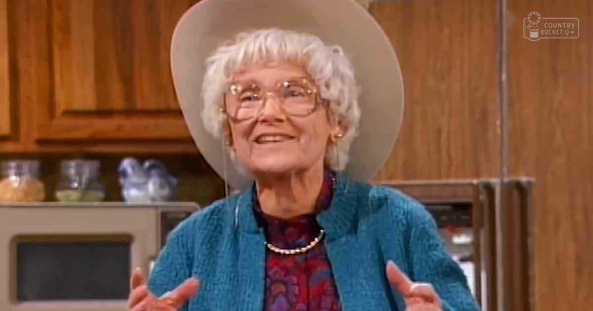 An older adult female wearing a blue textured jacket over a patterned shirt with a high neckline and long sleeves, pearl necklace, in a kitchen setting with wooden cabinets, microwave, and glass jars.