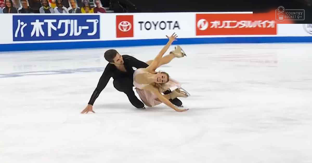 A figure skating pair performs at an indoor ice rink with spectators and advertising boards in the background, under artificial lighting.