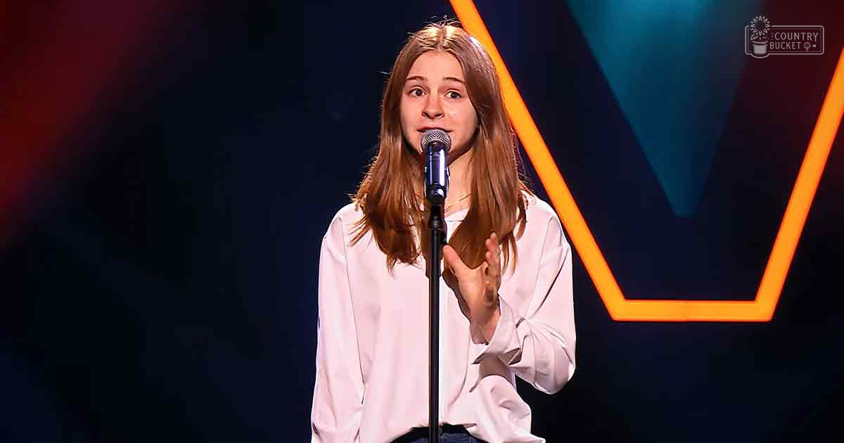A young adult female in a light pink blouse performs on stage with bright orange geometric shapes and professional lighting in the background.