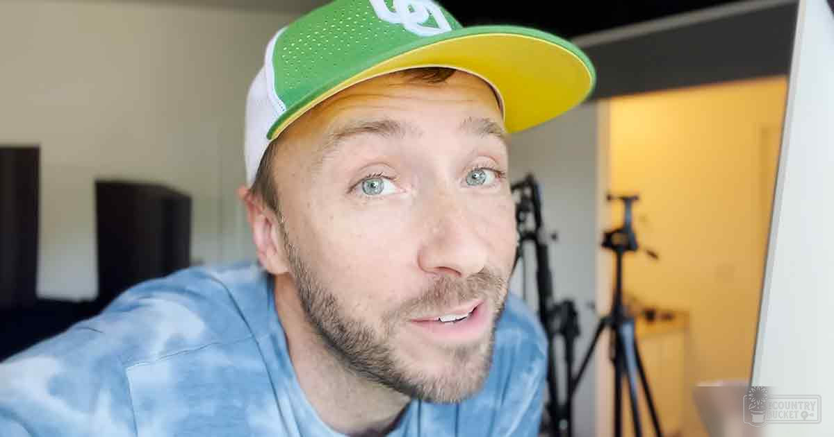 A young adult male wearing a blue tie-dye t-shirt, green baseball cap, and standing indoors near a door frame with natural lighting in the background.
