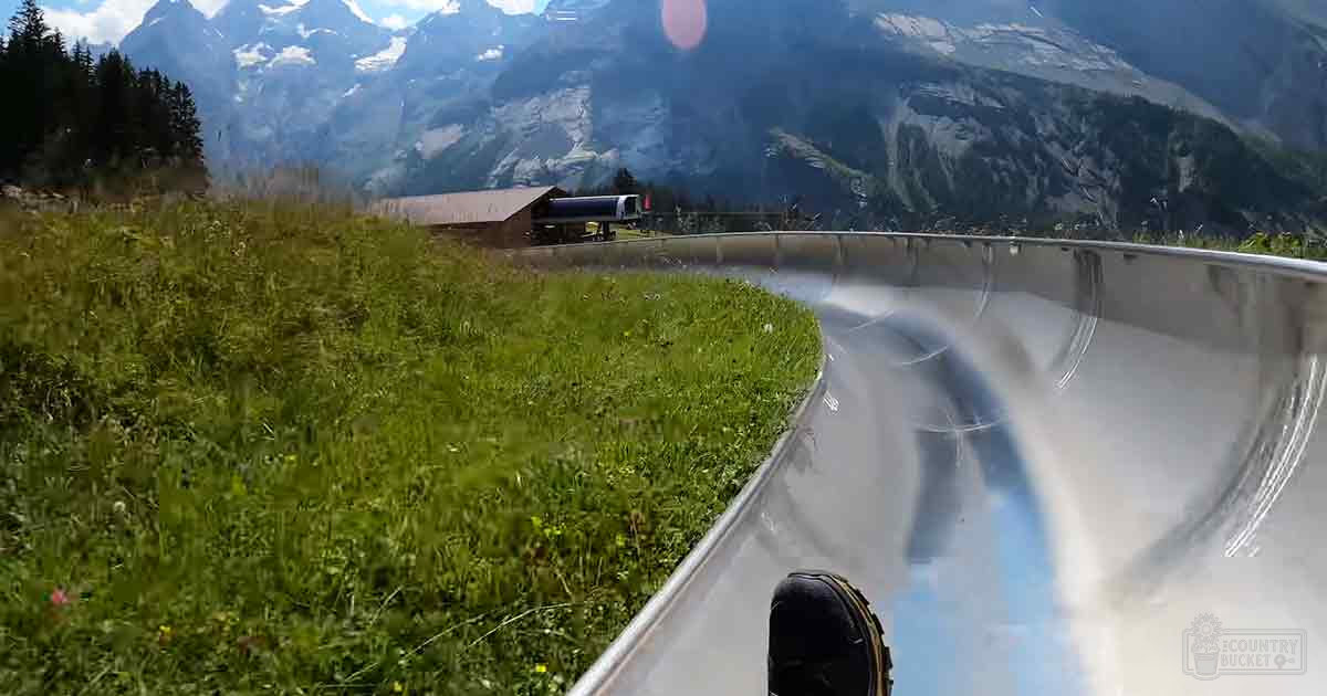 An alpine setting with a metal luge track, grassy fields, snow-capped mountains, and clear skies