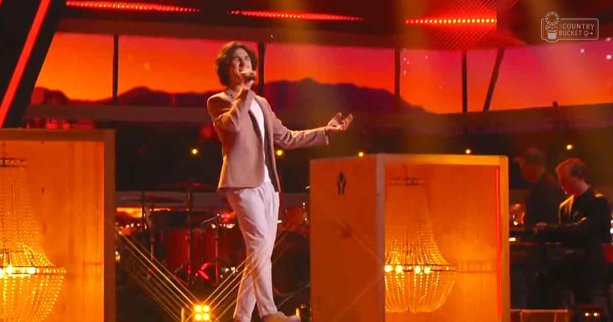 A young adult male singer performs on stage at a concert venue, wearing a light-colored blazer and white pants with red LED lights illuminating the backdrop with an orange sky with mountain silhouettes.