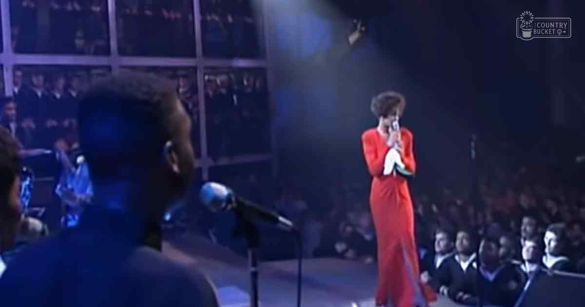An adult female performer in a bright red jumpsuit holding a microphone on stage with an audience seated in rows at an indoor concert venue under artificial stage lighting.