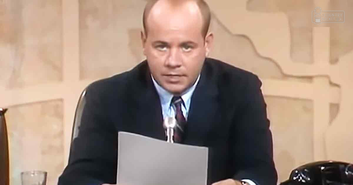 A man, likely an adult male in a professional setting, sits at what appears to be a news desk. He wears a dark suit jacket with a light blue dress shirt and patterned tie.