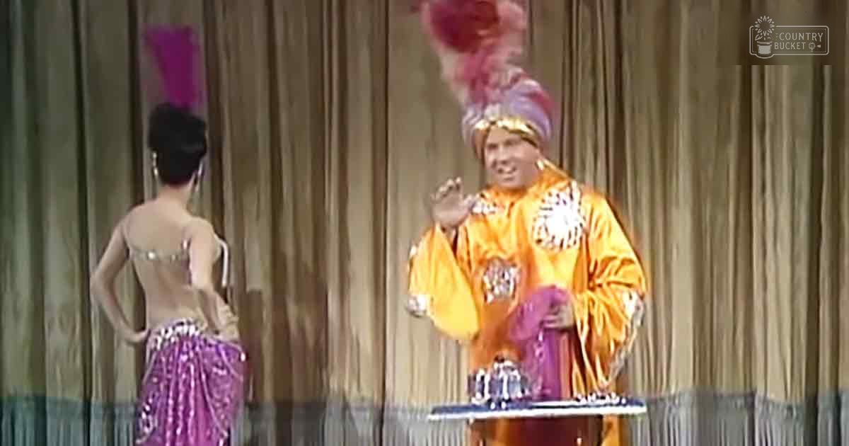 A theatrical performance with two performers; the male on the right wears a bright orange robe and large headdress, while the female on the left dons an exotic outfit in shiny sequined fabric. The setting is indoors, likely a stage.