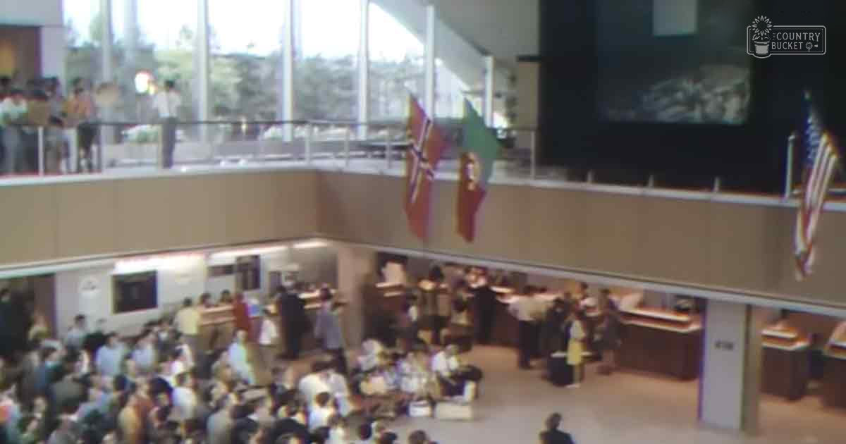 A bustling indoor public event with multiple levels, with adults in casual attire and business casual, flags prominently displayed, structured layout suggesting government building or institution.